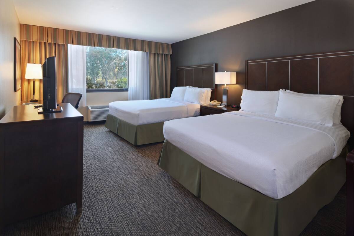 A hotel room with two beds, a desk, a TV, and a window with curtains.