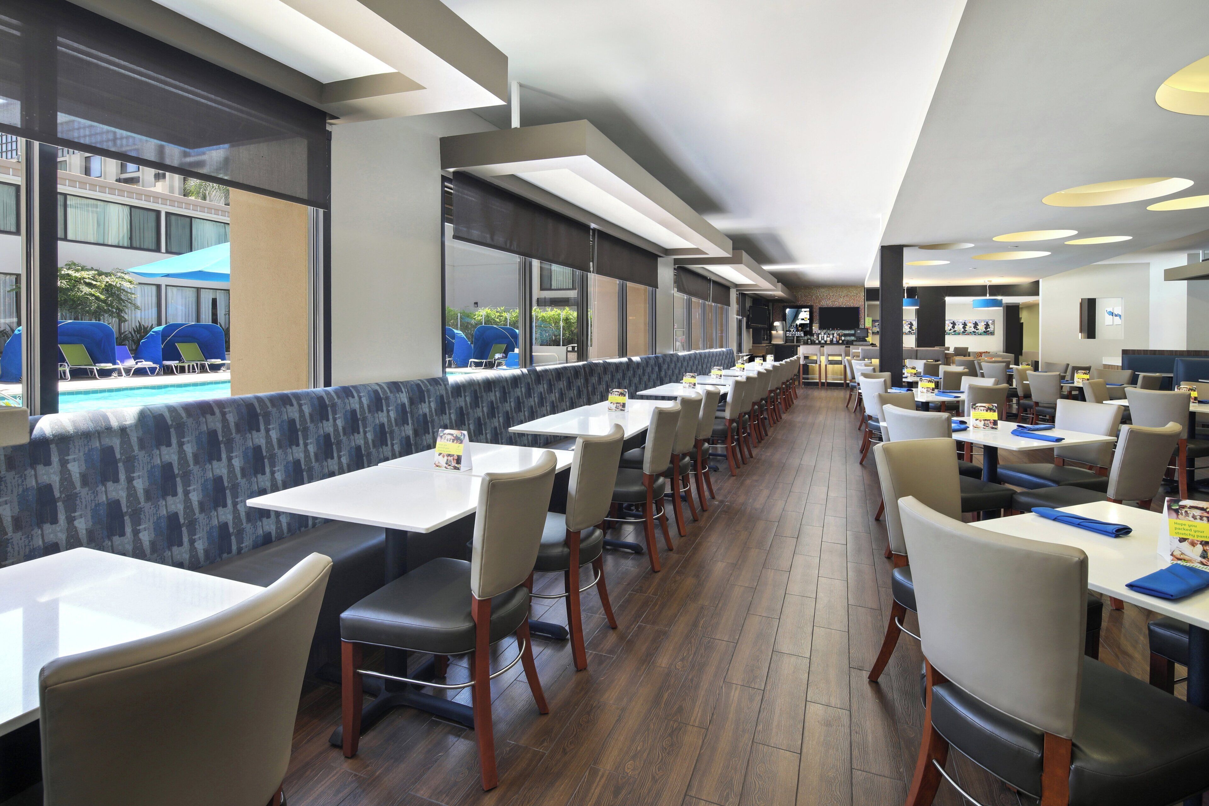 Un restaurante moderno con mesas y sillas largas, ventanas amplias y vista a una piscina.