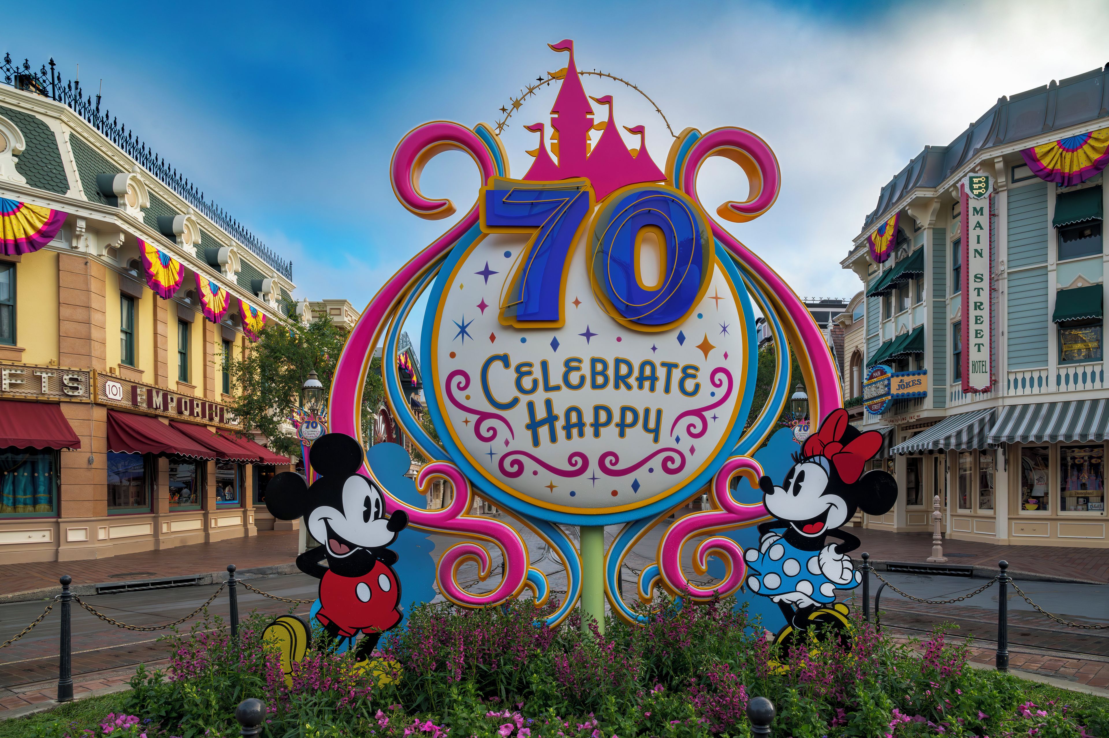A colorful 70th anniversary celebration sign with Mickey and Minnie Mouse, surrounded by flowers and classic architecture.