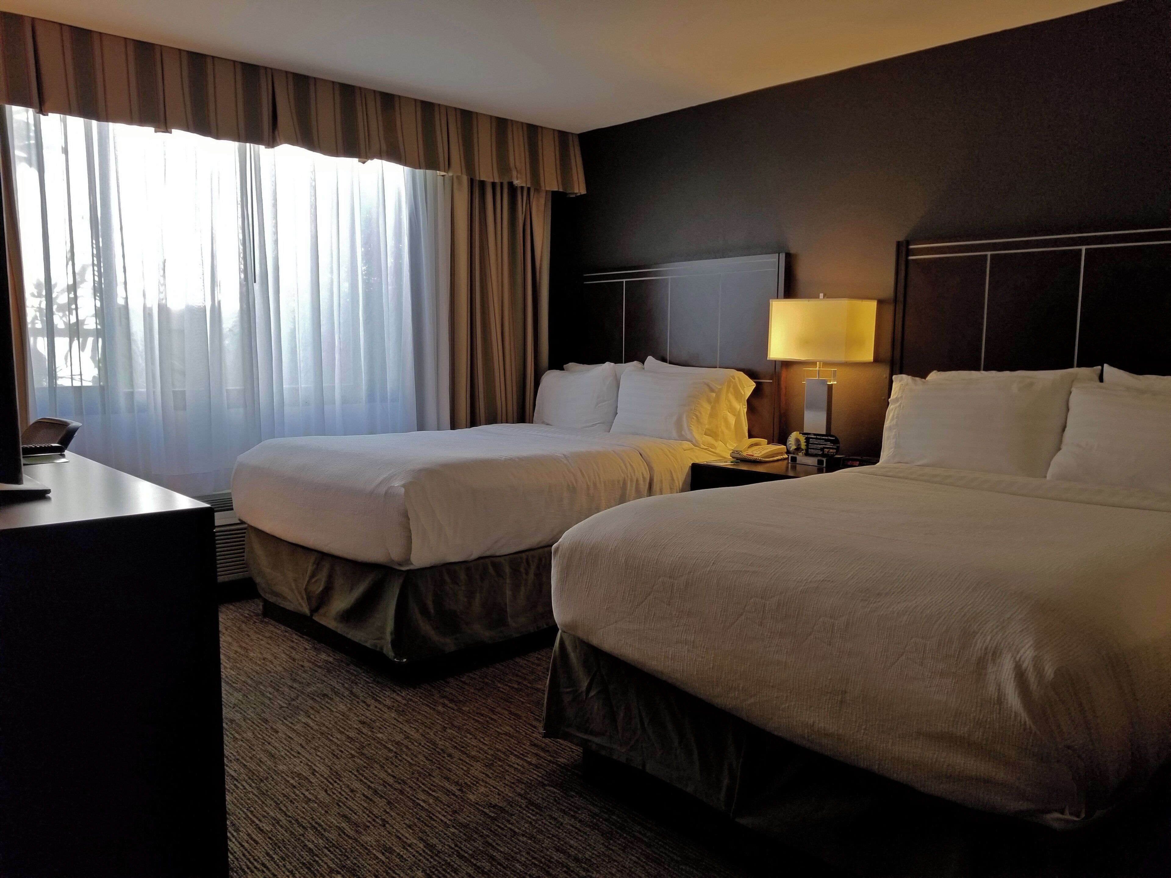 A hotel room with two beds, a desk, a lamp, and a window with curtains.
