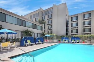 Zona de piscina de un hotel con tumbonas y sombrillas, rodeada por un edificio con varias ventanas y balcones.