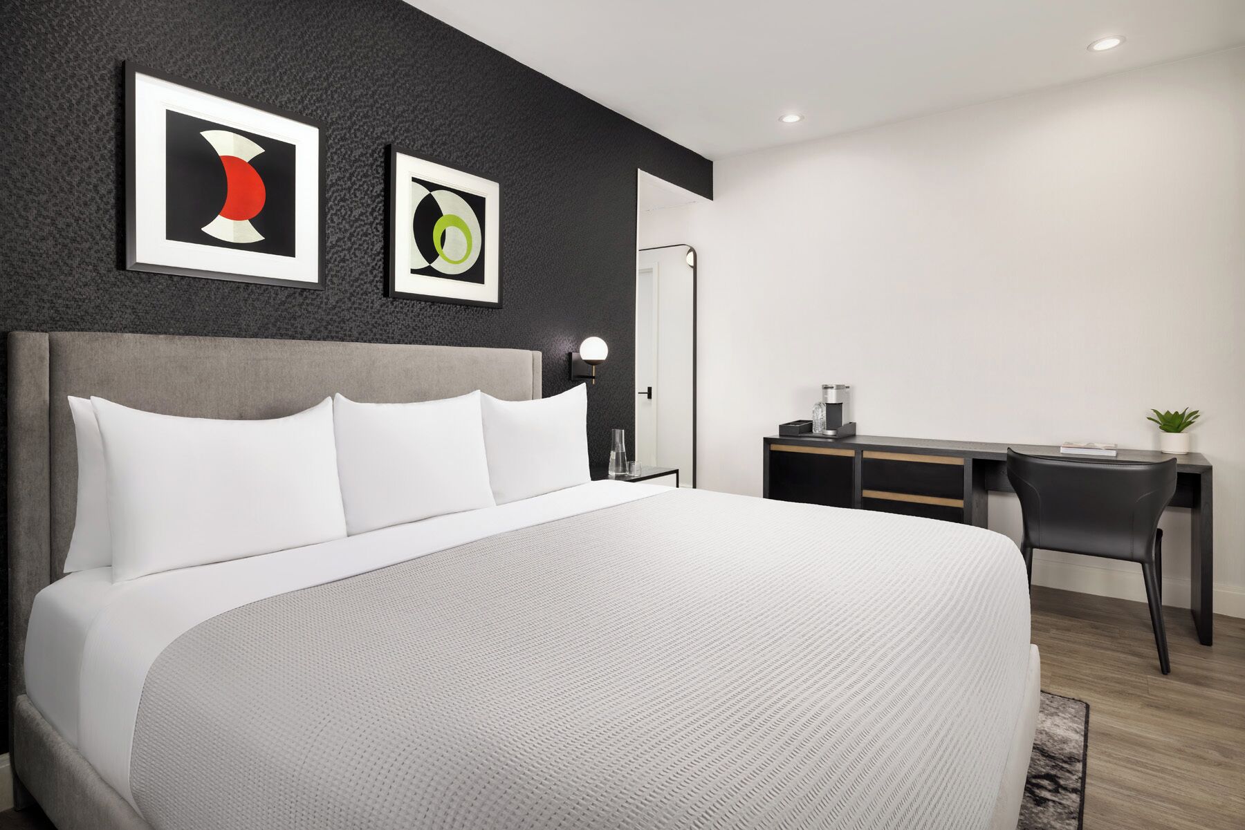 A modern hotel room with a large bed, a desk, and two abstract paintings on the wall.