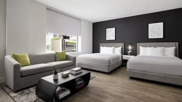 A modern hotel room with a grey sofa, two single beds, a small coffee table, and a view of the city through large windows.