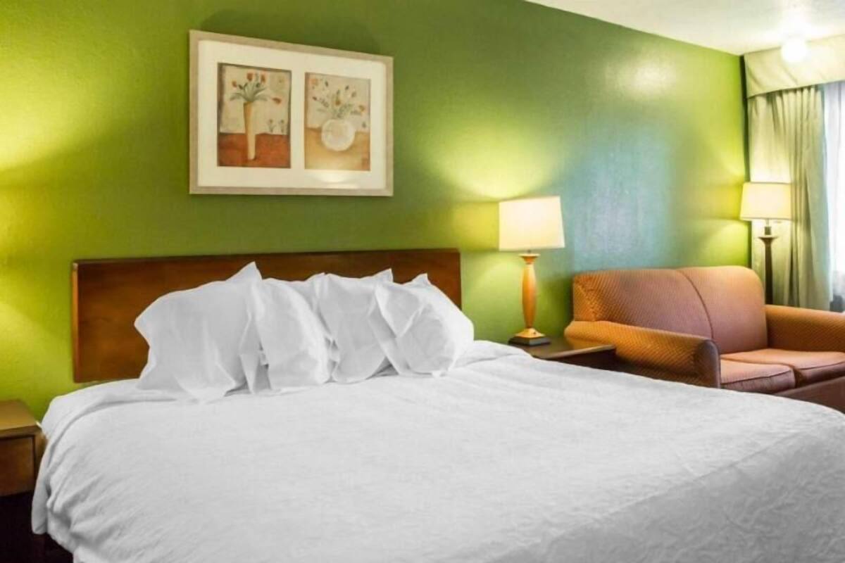 A hotel room with a bed, a nightstand, a lamp, a chair, and a painting on the wall.