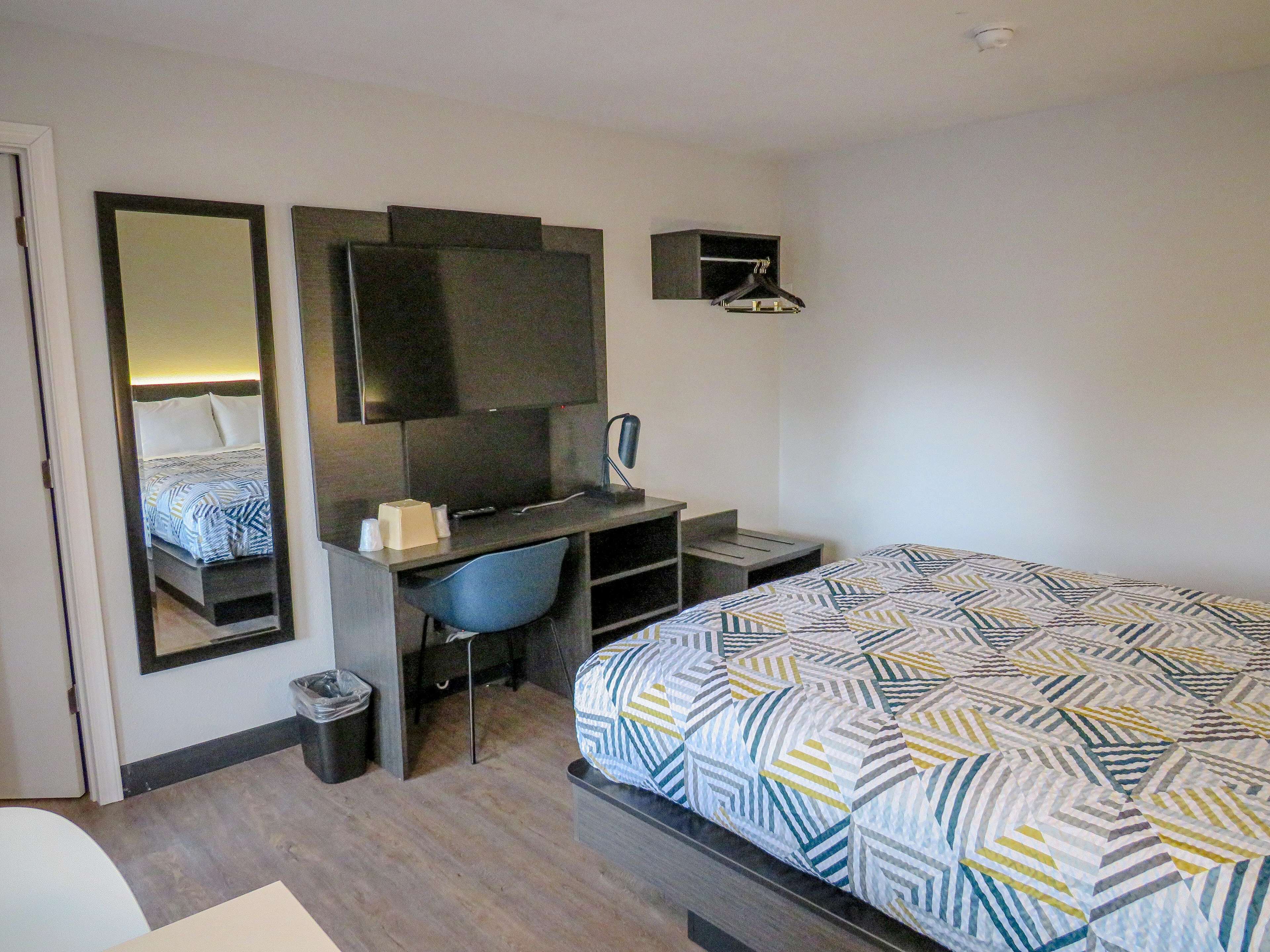A hotel room with a bed, a desk with a chair, a mirror, a television, and a small shelf.