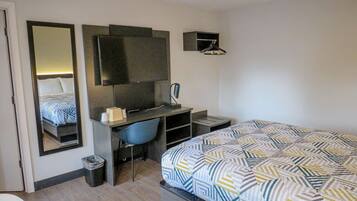 A hotel room with a bed, a desk with a chair, a mirror, a television, and a small shelf.