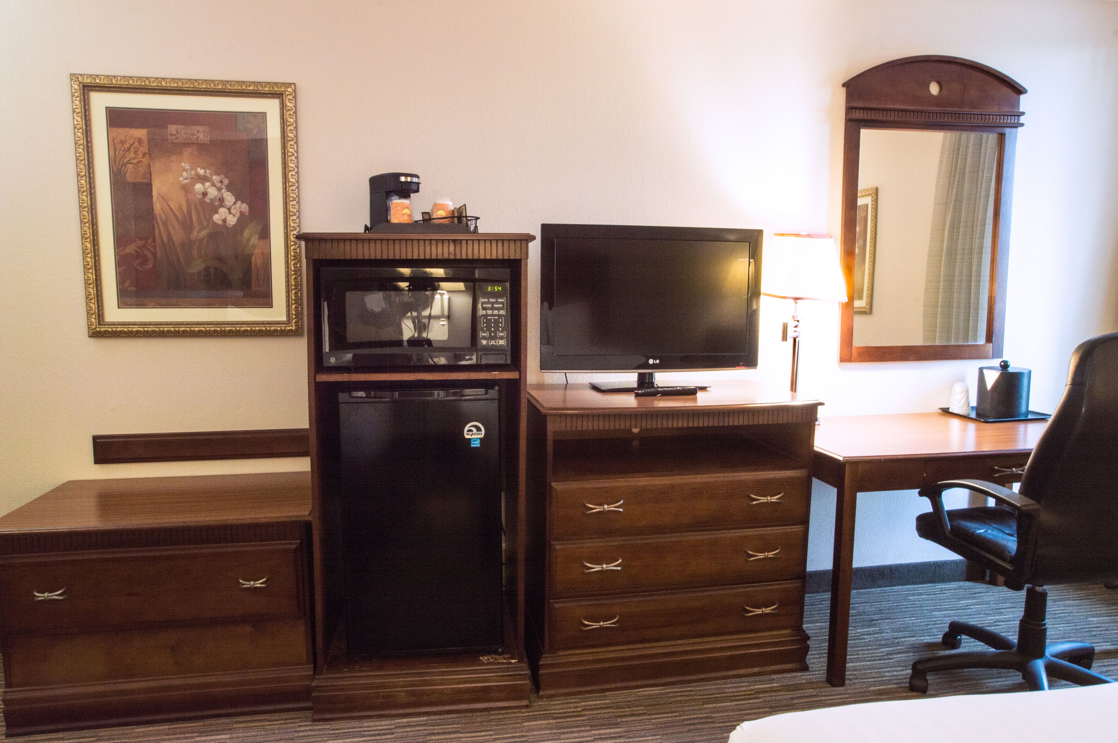 A hotel room with a wooden desk, a television, a refrigerator, a chair, and a framed picture on the wall.