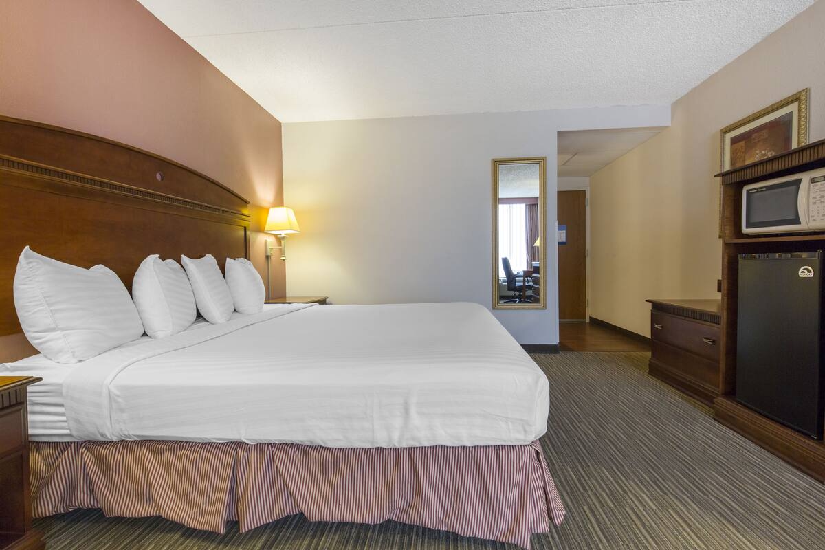 A hotel room with a large bed, a nightstand, a lamp, a dresser, a microwave, and a mirror.