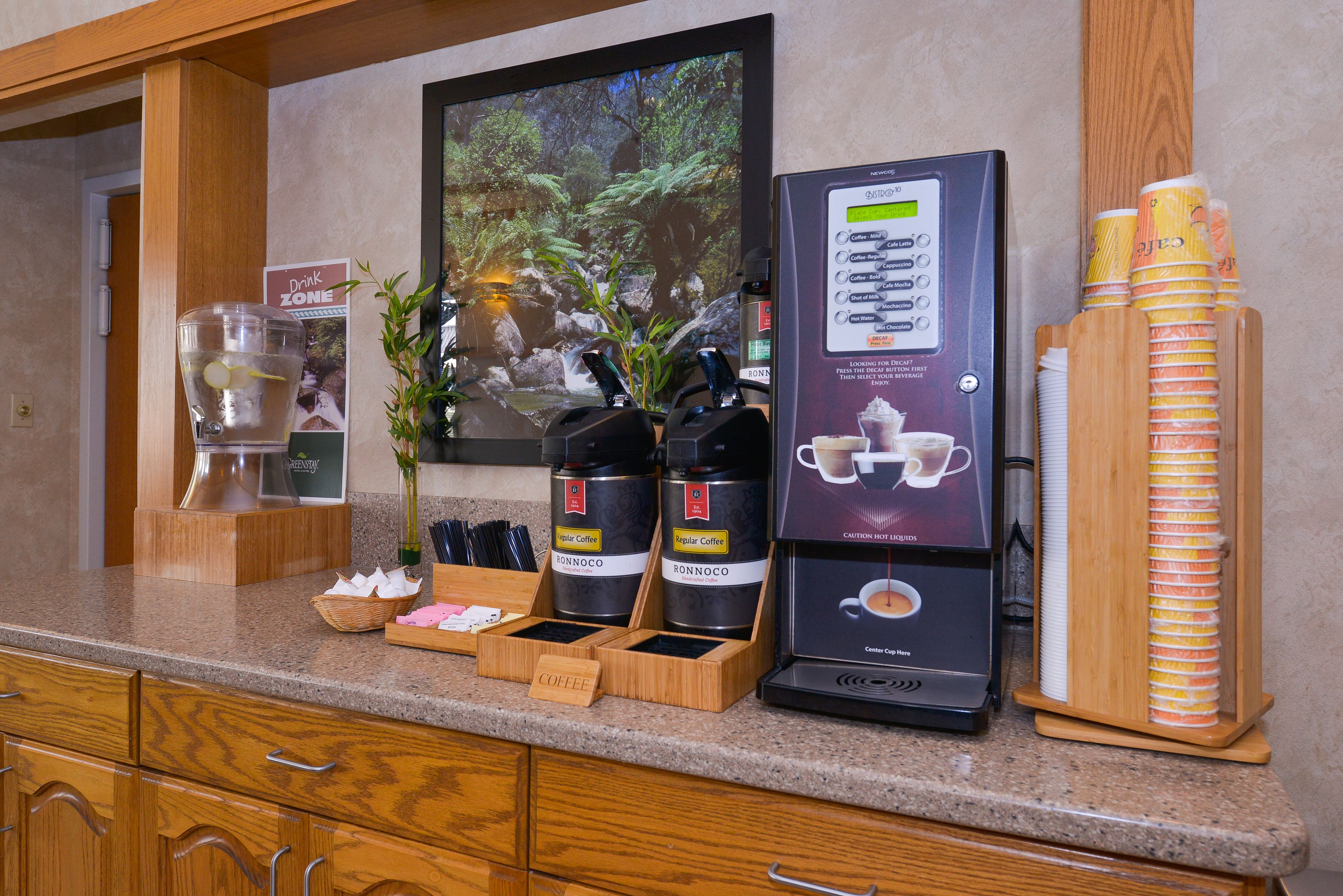 A coffee station with a coffee machine, two coffee dispensers, a drink sign, and a wall-mounted screen displaying a nature scene.