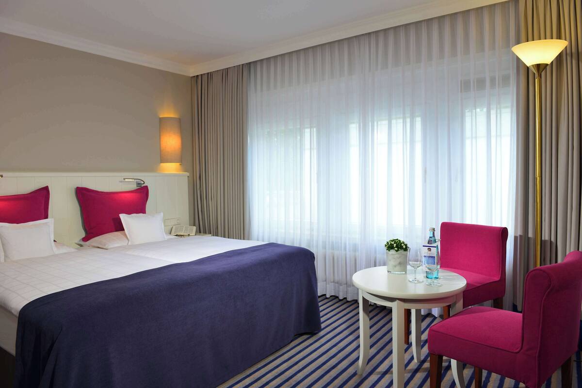 Une chambre d’hôtel avec un lit, une table ronde entourée de chaises, une lampe et une fenêtre avec des rideaux.