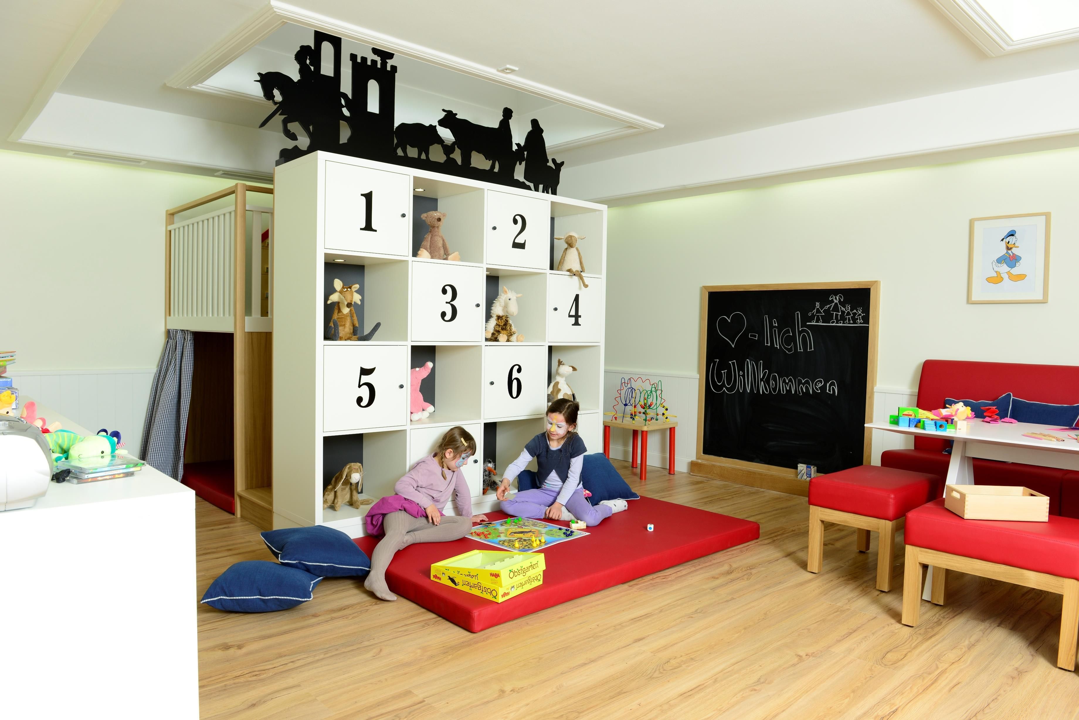 Una sala de juegos infantil con un mueble de almacenamiento con motivos numéricos, un tapete de juego rojo y una pizarra con la palabra 'Welcome'.