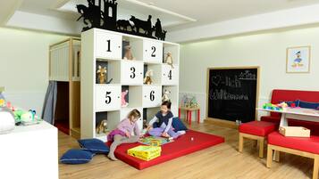 Una sala de juegos infantil con un mueble de almacenamiento con motivos numéricos, un tapete de juego rojo y una pizarra con la palabra 'Welcome'.