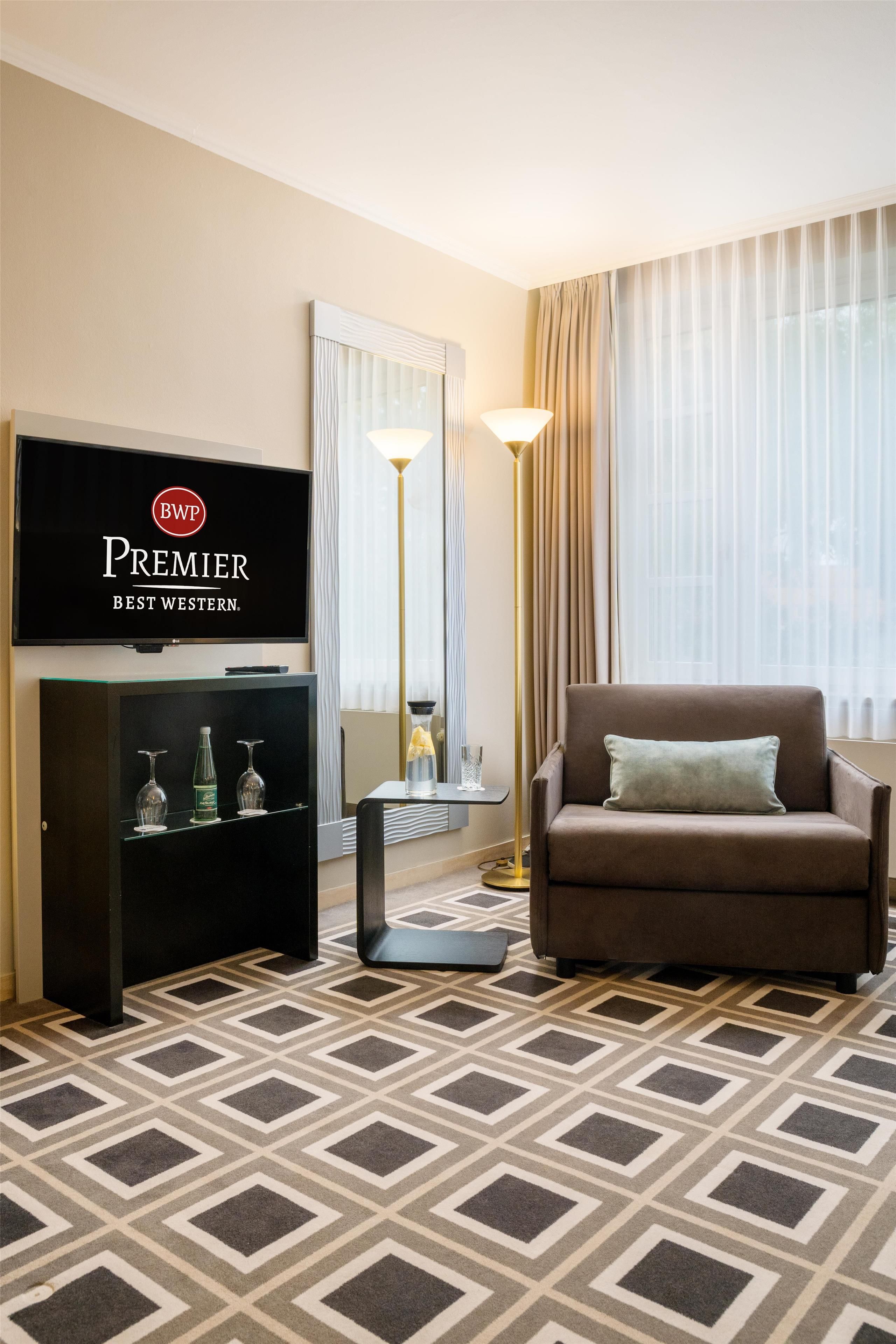 Habitación de hotel con una televisión mostrando la marca 'Premier Best Western', un sofá marrón con un cojín, una alfombra estampada y una lámpara de pie.