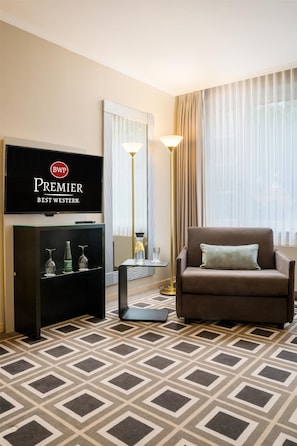 A hotel room with a television displaying 'Premier Best Western' branding, a brown sofa with a cushion, a patterned carpet, and a floor lamp. - Best Western Premier Parkhotel Kronsberg (Hannover)