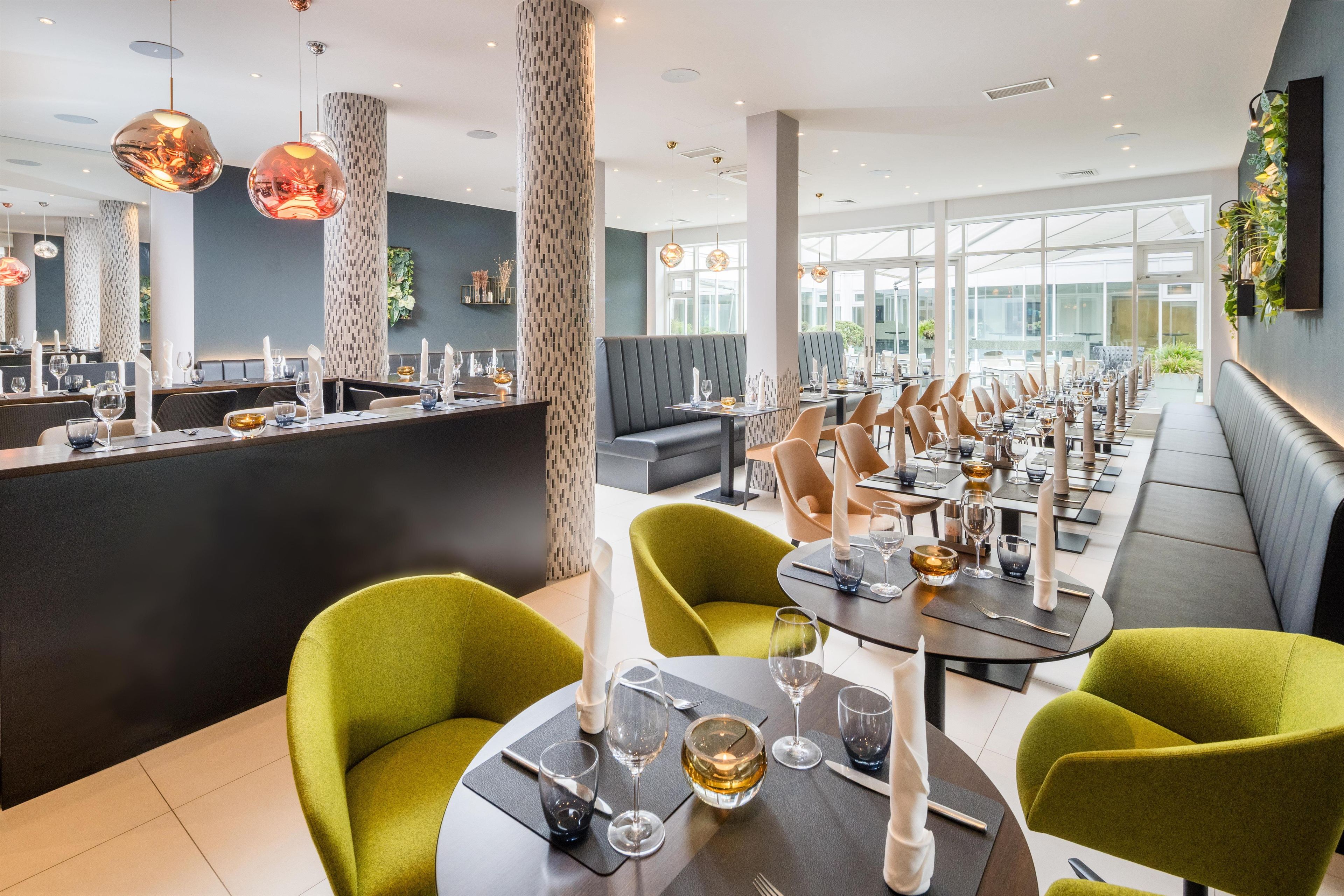 A modern restaurant interior with high ceilings, patterned columns, and a long dining area with green upholstered chairs and tables.