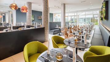 A modern restaurant interior with high ceilings, patterned columns, and a long dining area with green upholstered chairs and tables.