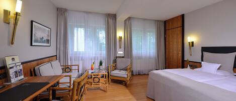A hotel room with a bed, a desk with a chair, a small table with a chair, and a window with curtains.