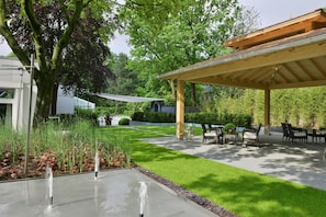 Een overdekt terras met een pergola, een waterpartij en een verzorgd gazon.
