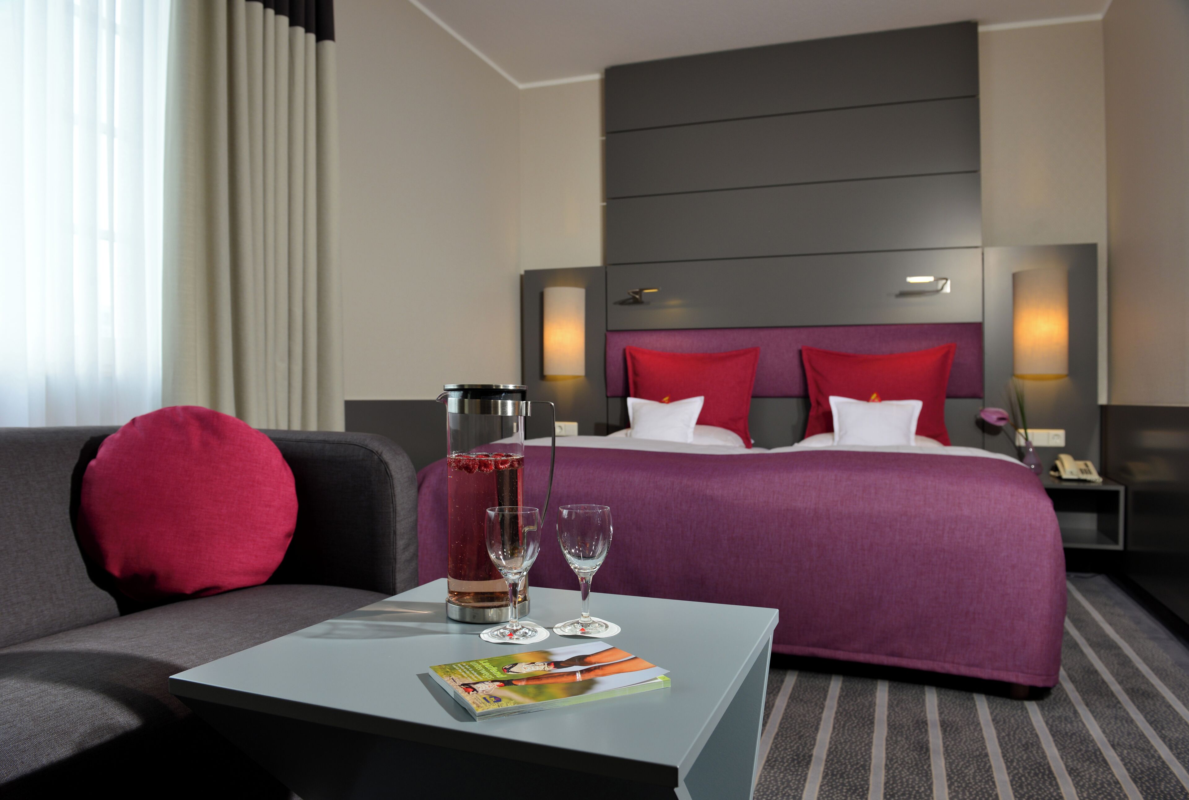A hotel room with a bed, a nightstand, a coffee maker, a table with a magazine, and a sofa.