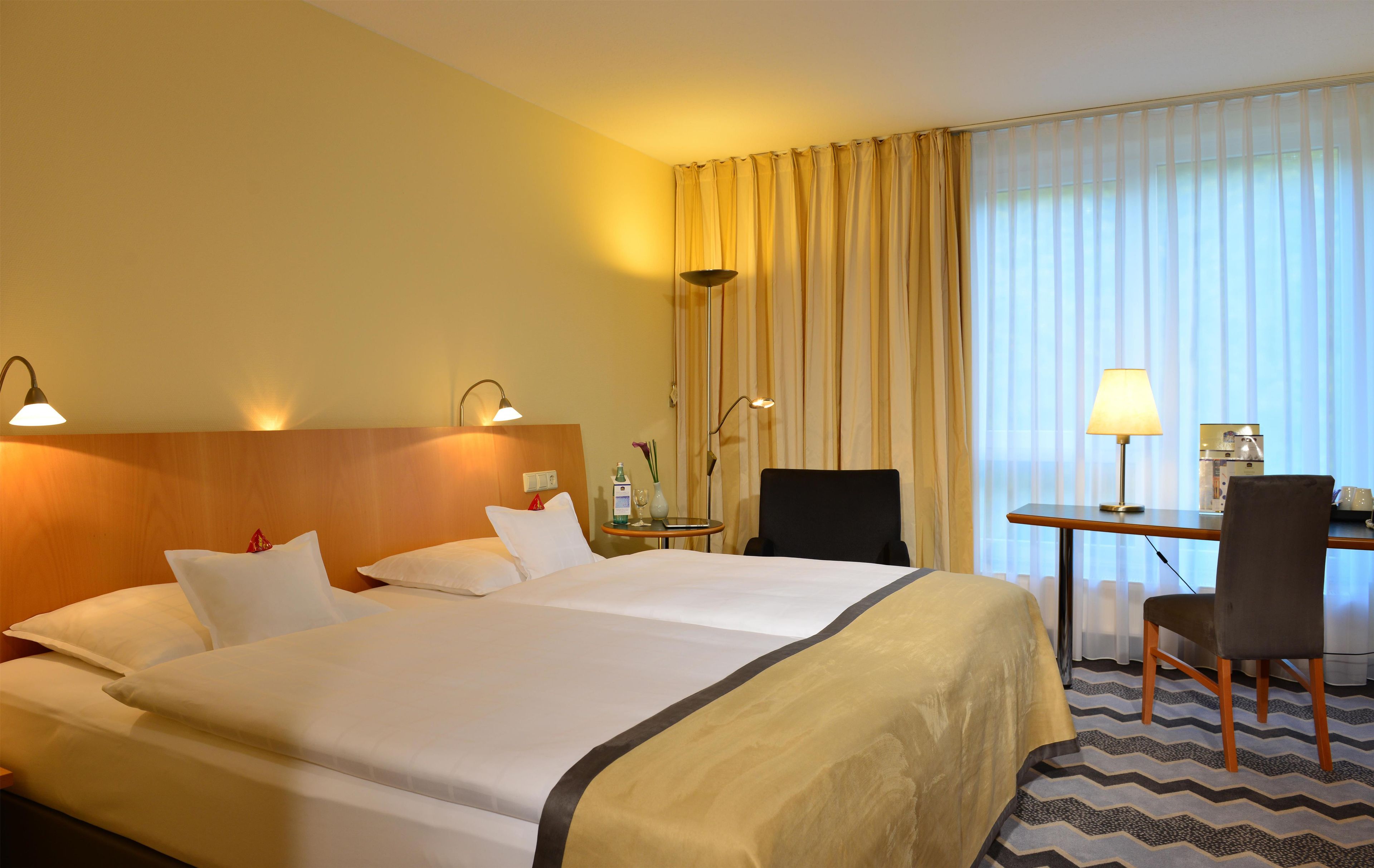 Ein Hotelzimmer mit Doppelbett, Nachttischlampen, einem Schreibtisch mit Stuhl und einem großen Fenster mit Vorhängen.