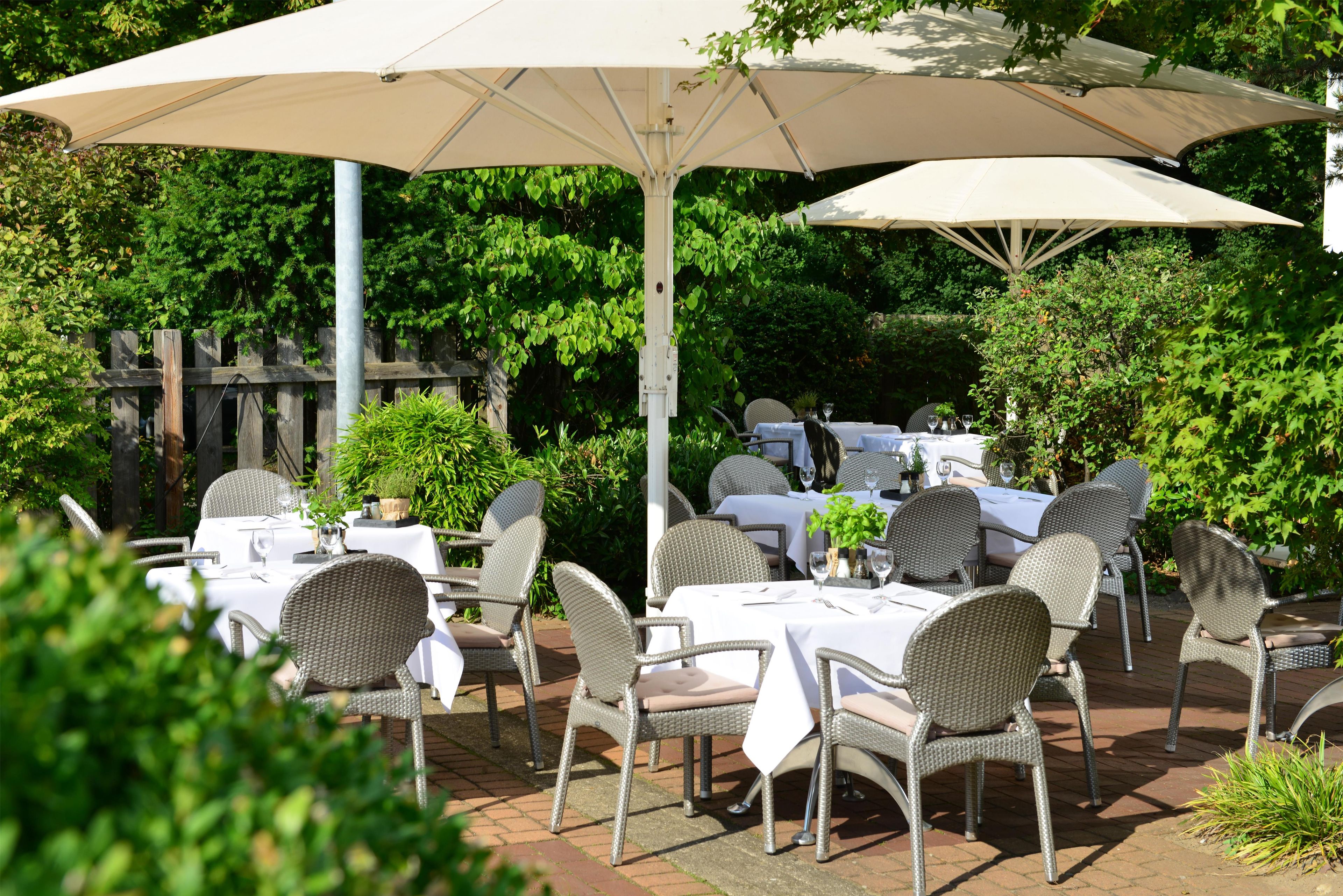 Eine Außenpatio mit Tischen und Stühlen, beschattet durch große Sonnenschirme, umgeben von viel Grün.