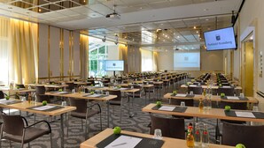 Een vergaderzaal met rijen tafels, stoelen en een groot scherm waarop 'Parkhotel Kronberg' staat.