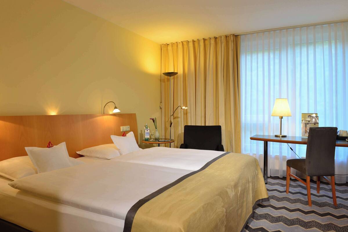 Une chambre d’hôtel avec un grand lit, deux lampes de chevet, un bureau avec une chaise et une grande fenêtre avec des rideaux.