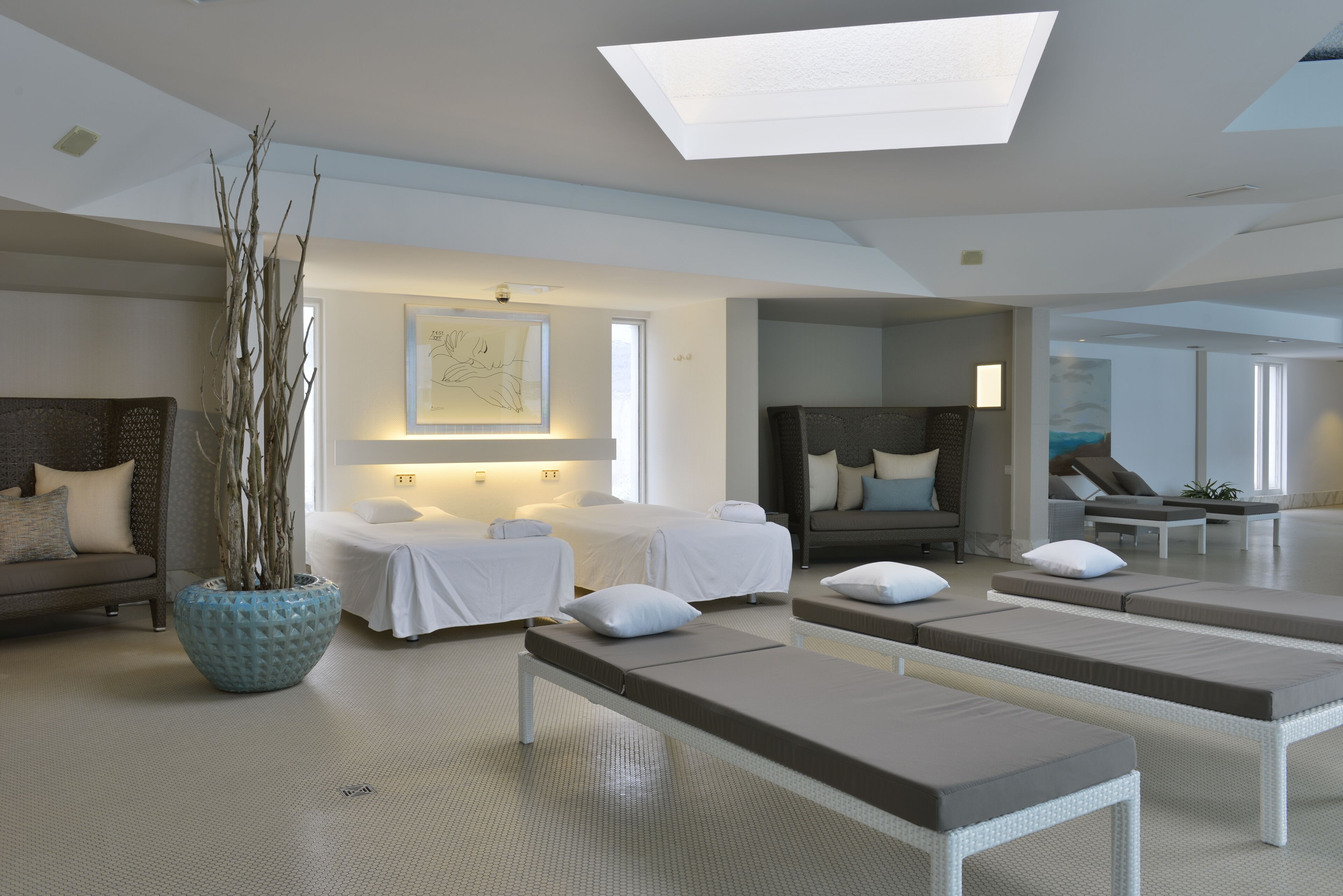 A modern spa room with a large skylight, a massage table, a lounge area, and a decorative plant.