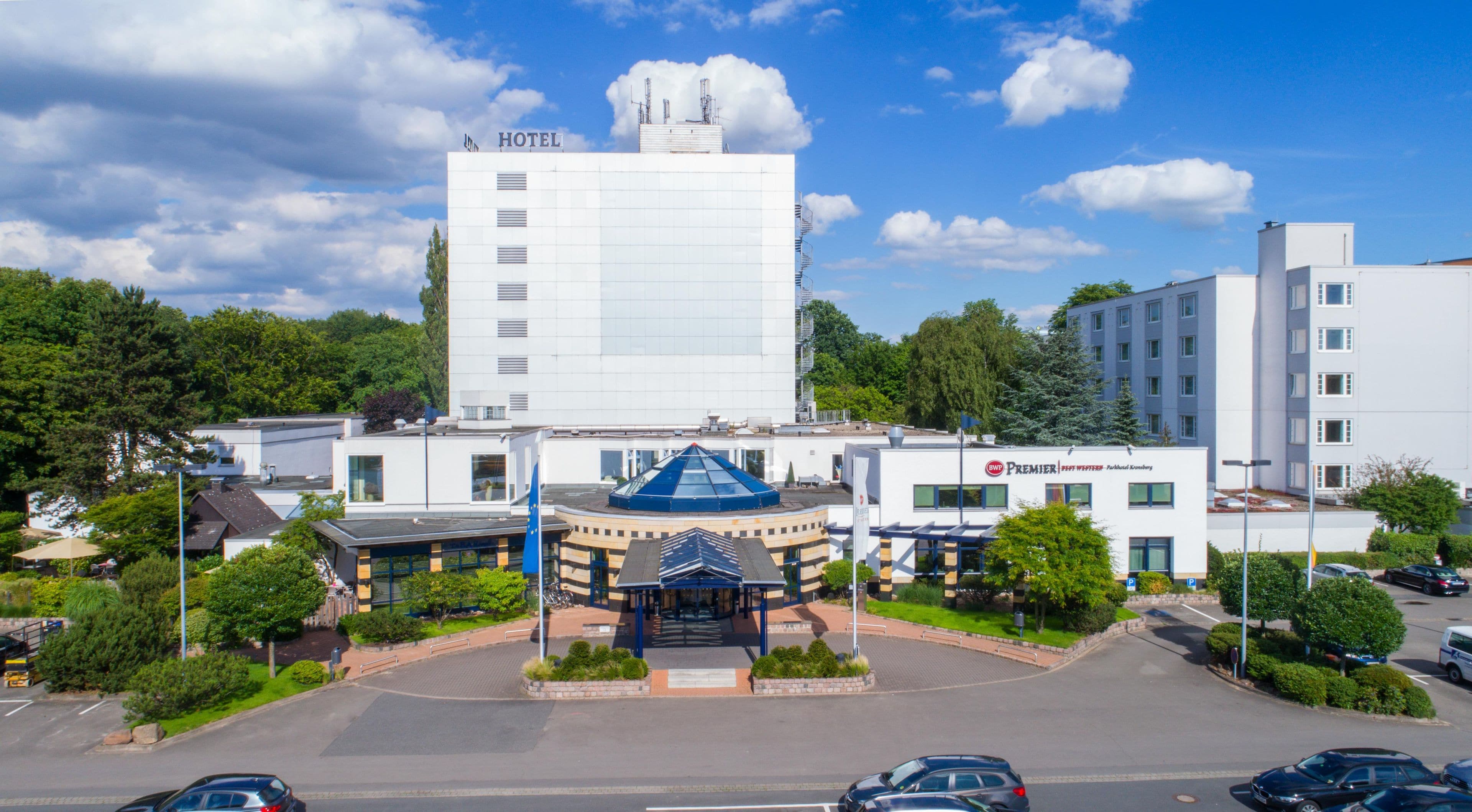 Ein Hotel mit einer markanten weißen Fassade, einem zentralen Eingang mit einem blauen Dach und einem Parkplatz davor.