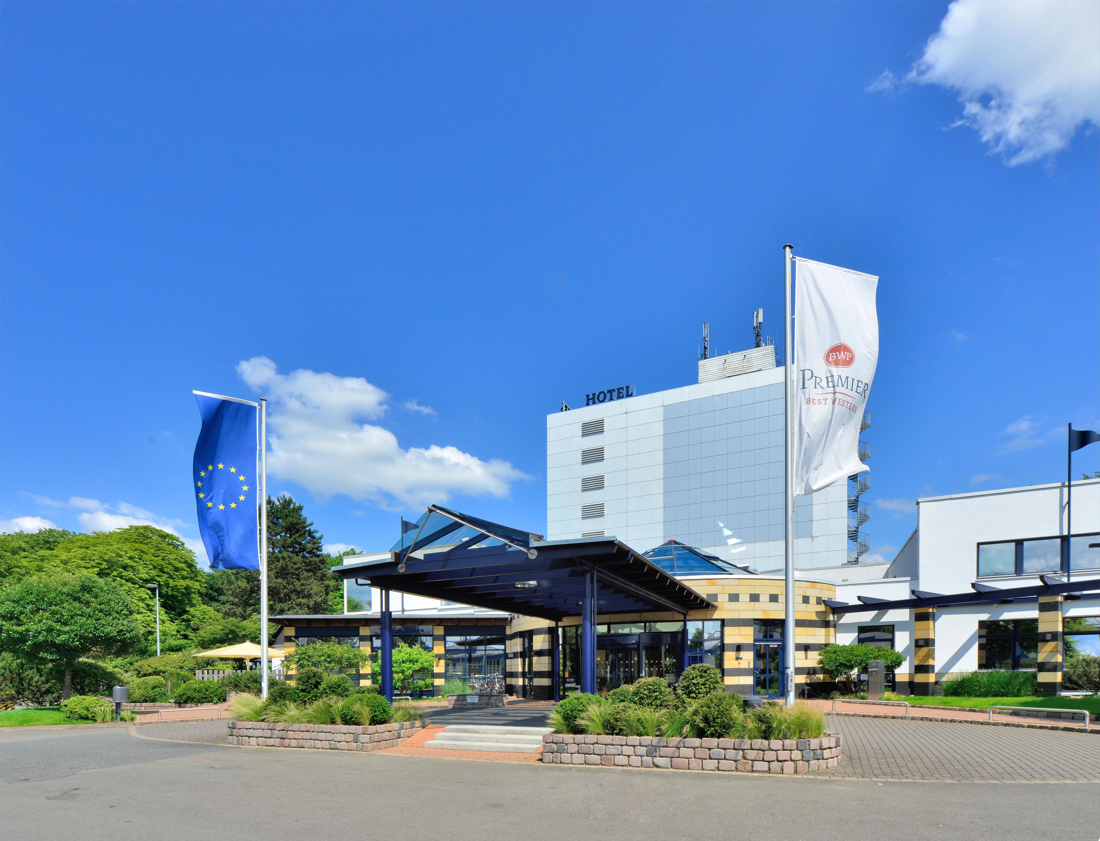 Ein Hotel mit moderner Architektur, das einen markanten Eingangsbereich mit einem überdachten Gang und einer Fahnenstange mit der Flagge der Europäischen Union zeigt.