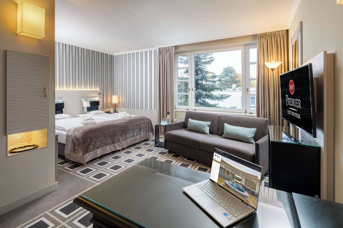Une chambre d’hôtel avec un lit, un canapé, une télévision et un ordinateur portable posé sur une table en verre.