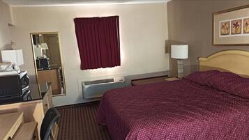 A hotel room with a bed, a desk with a computer, a mirror, a window with a red curtain, an air conditioning unit, and a framed picture on the wall.