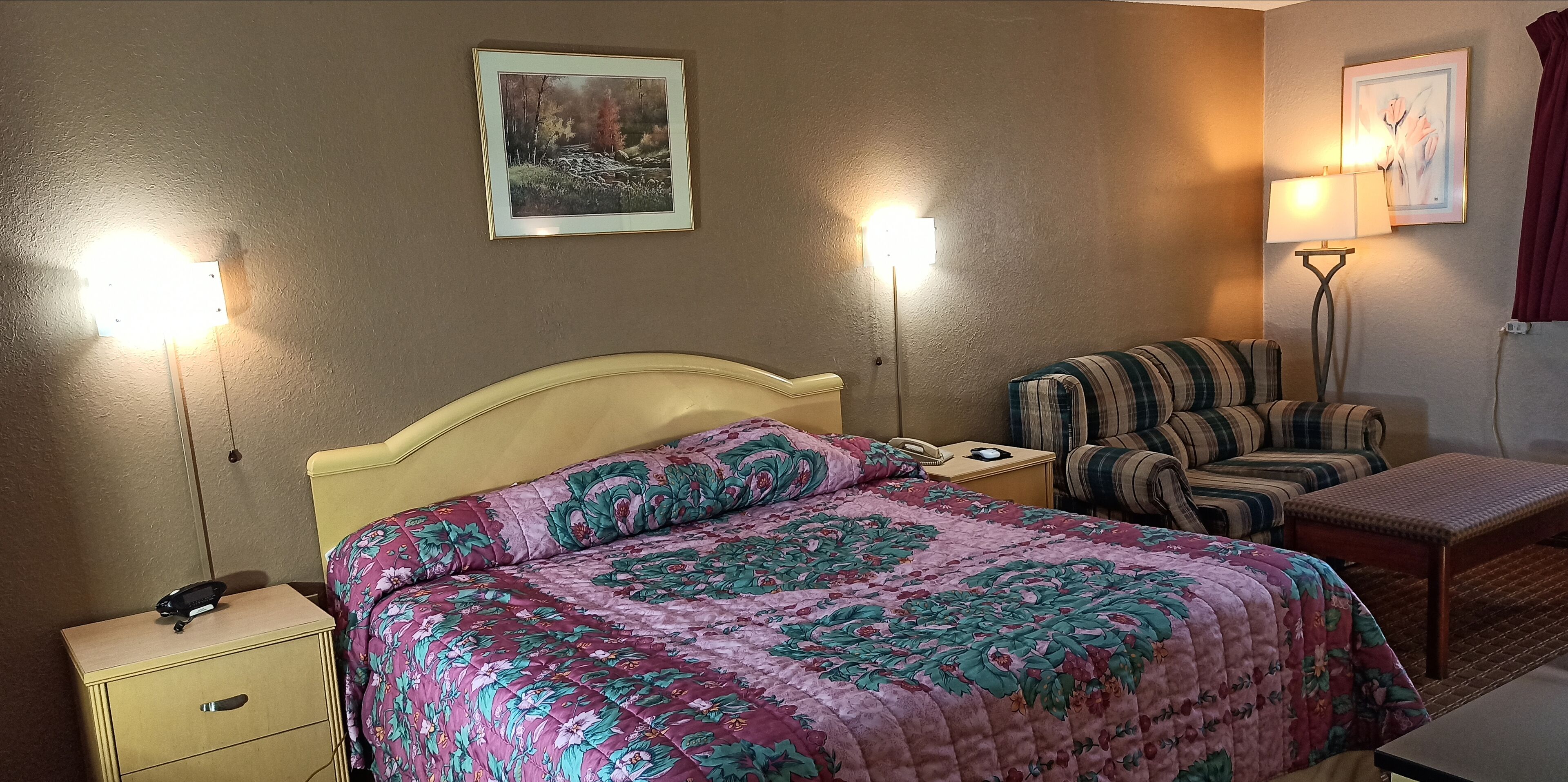 A hotel room with a bed, a chair, a bedside table, a lamp, and a painting on the wall.