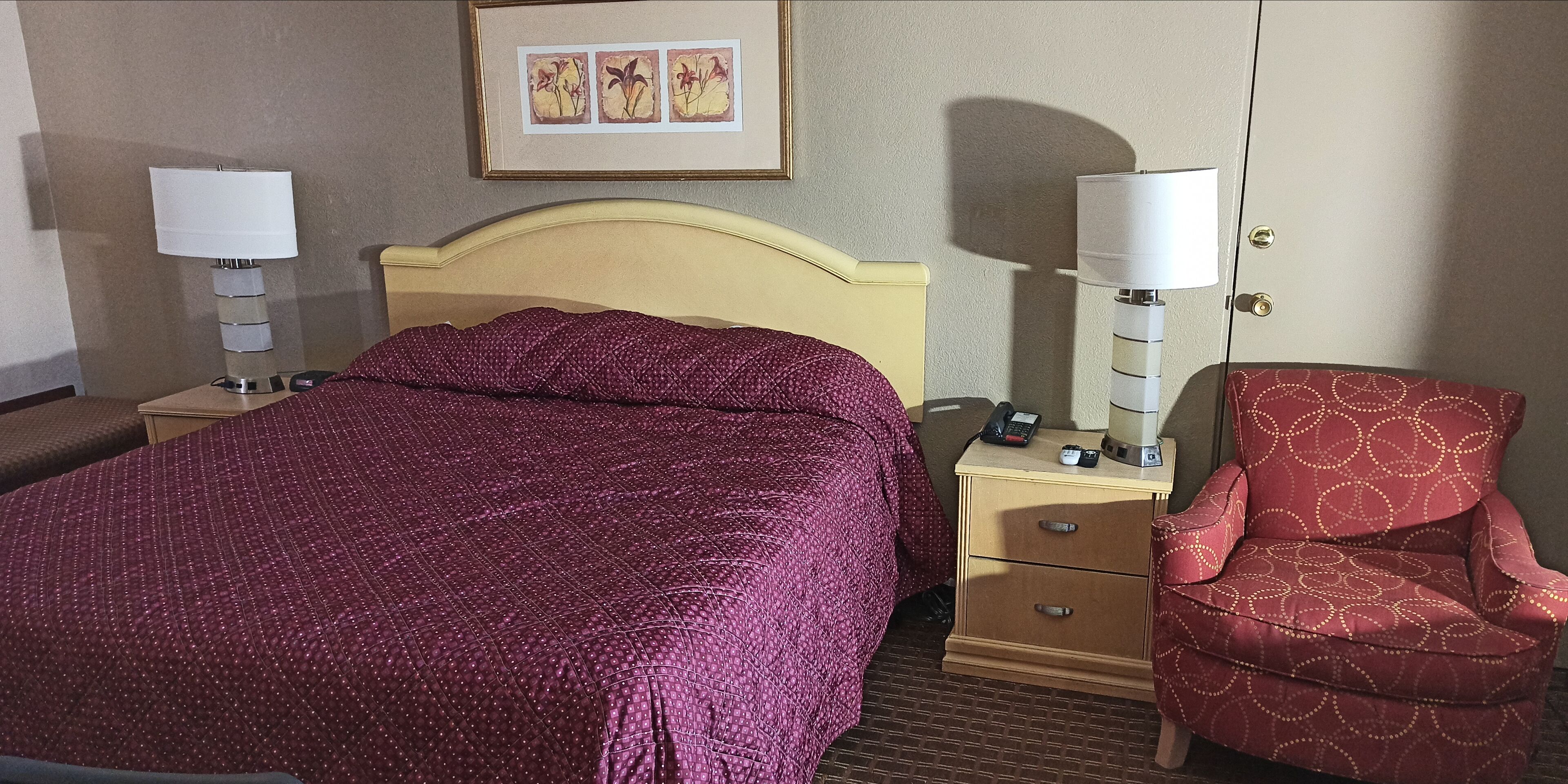 Een hotelkamer met een bed, nachtkastje, stoel, lamp en een fotokader aan de muur.
