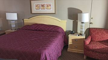 Een hotelkamer met een bed, nachtkastje, stoel, lamp en een fotokader aan de muur.