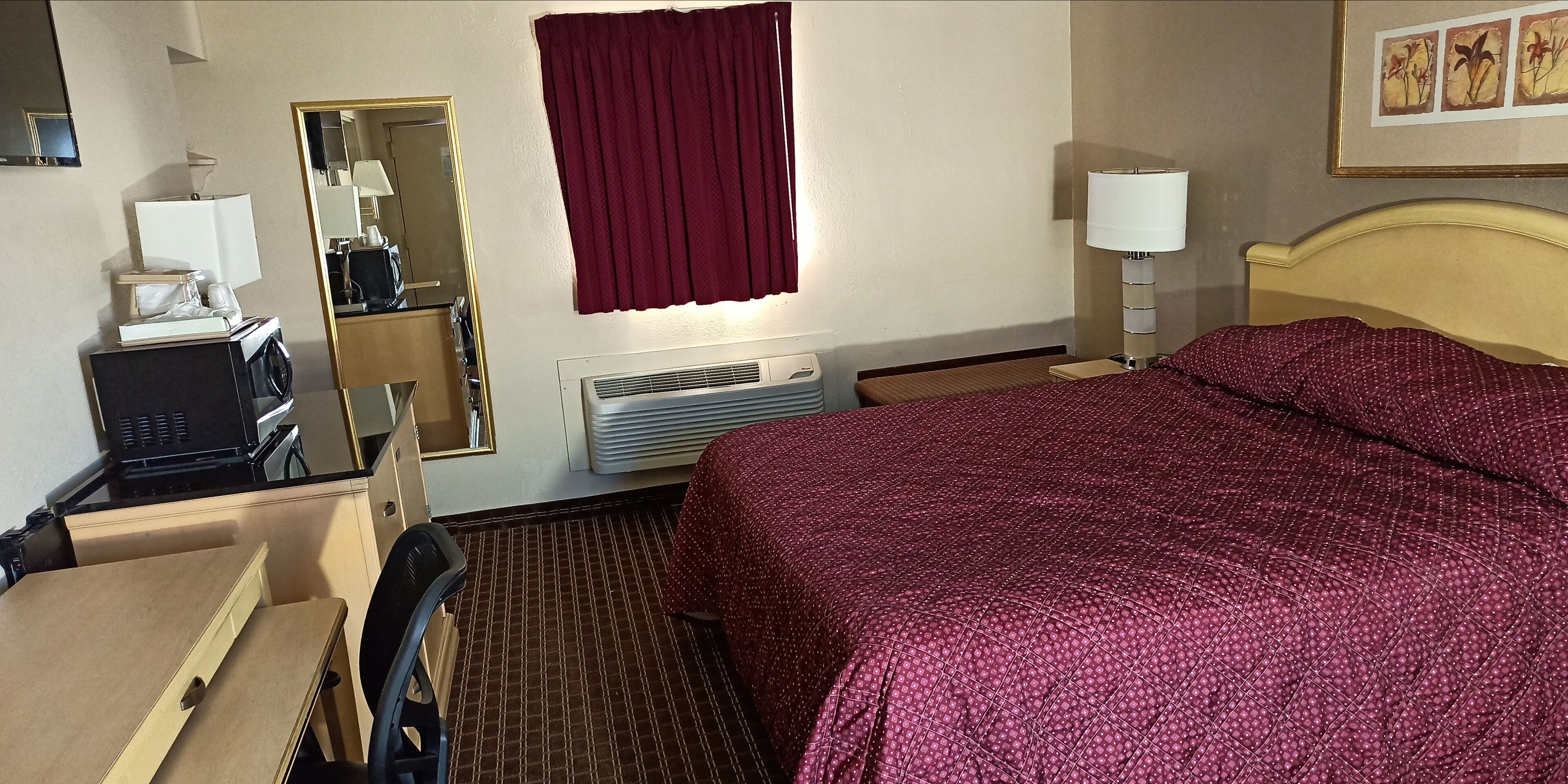 A hotel room with a bed, a desk, a chair, a mirror, a window with curtains, an air conditioning unit, and a lamp.