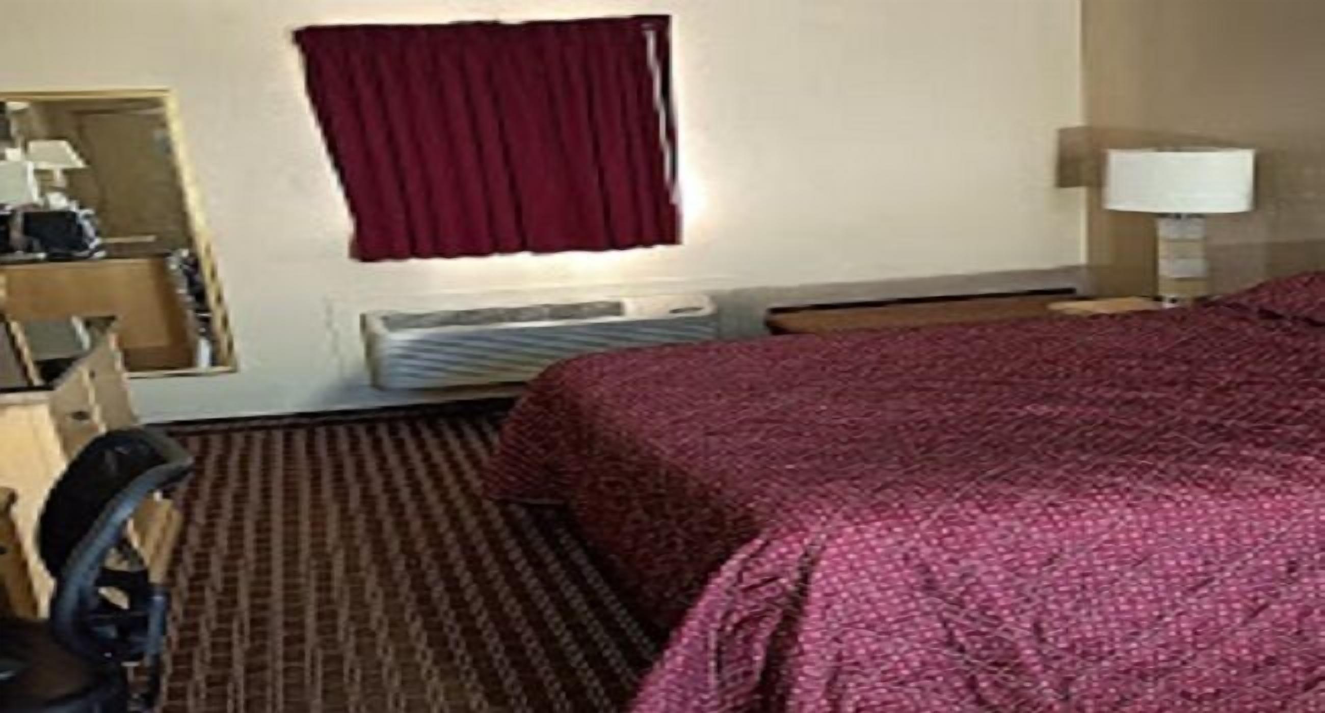 Une chambre d’hôtel avec un lit, un bureau et une chaise, une fenêtre avec un rideau rouge, une lampe et un radiateur.