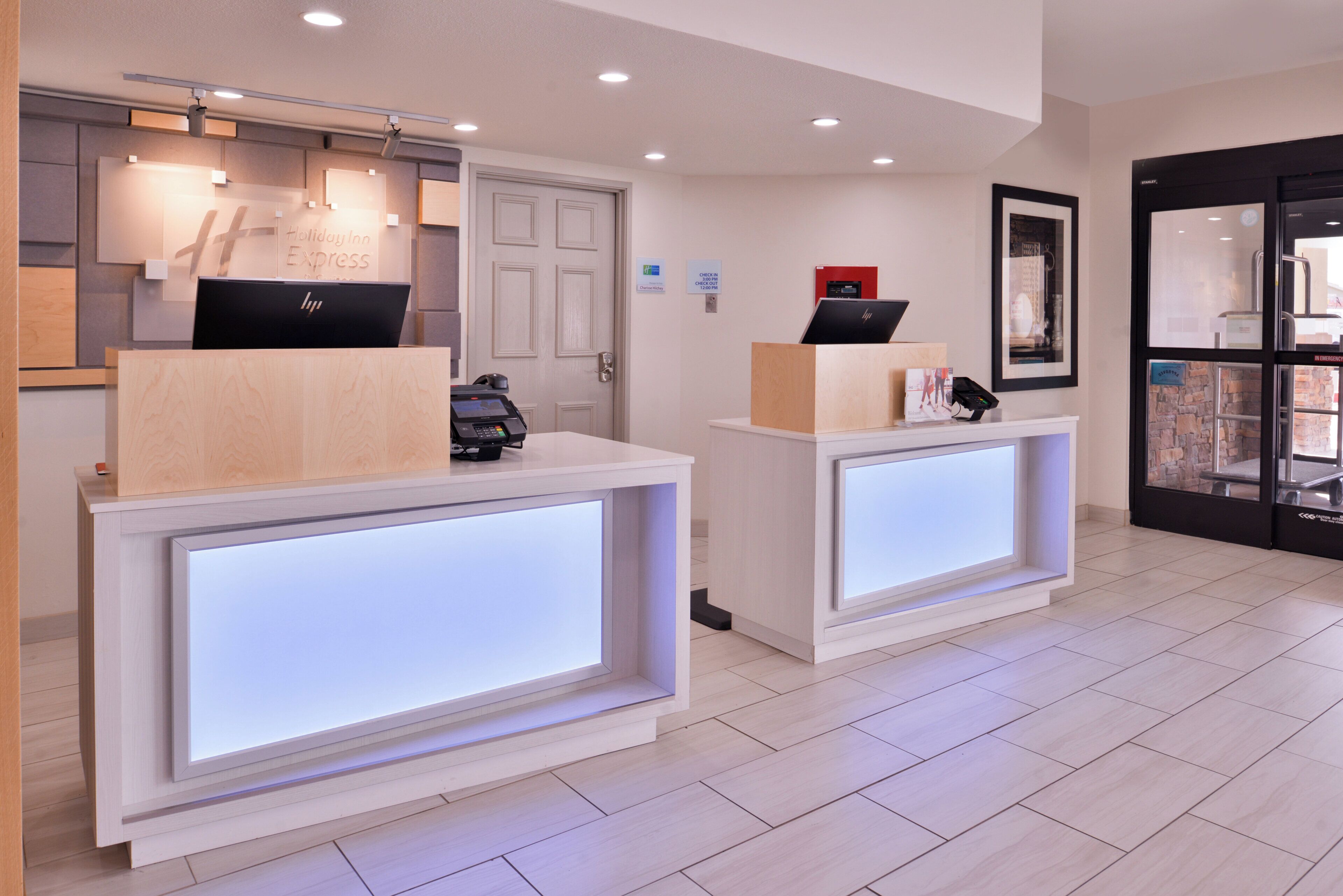 A hotel reception area with two reception desks, a digital display, a laptop, and a framed picture on the wall.