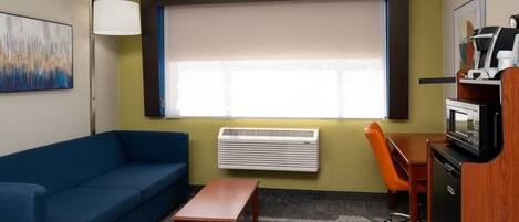 A hotel room with a blue sofa, a wooden table, a desk with a chair, a coffee machine, and a large window with blinds.
