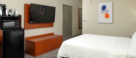 A hotel room with a bed, a TV mounted on a wooden panel, a microwave, a wooden dresser, and a framed artwork on the wall.
