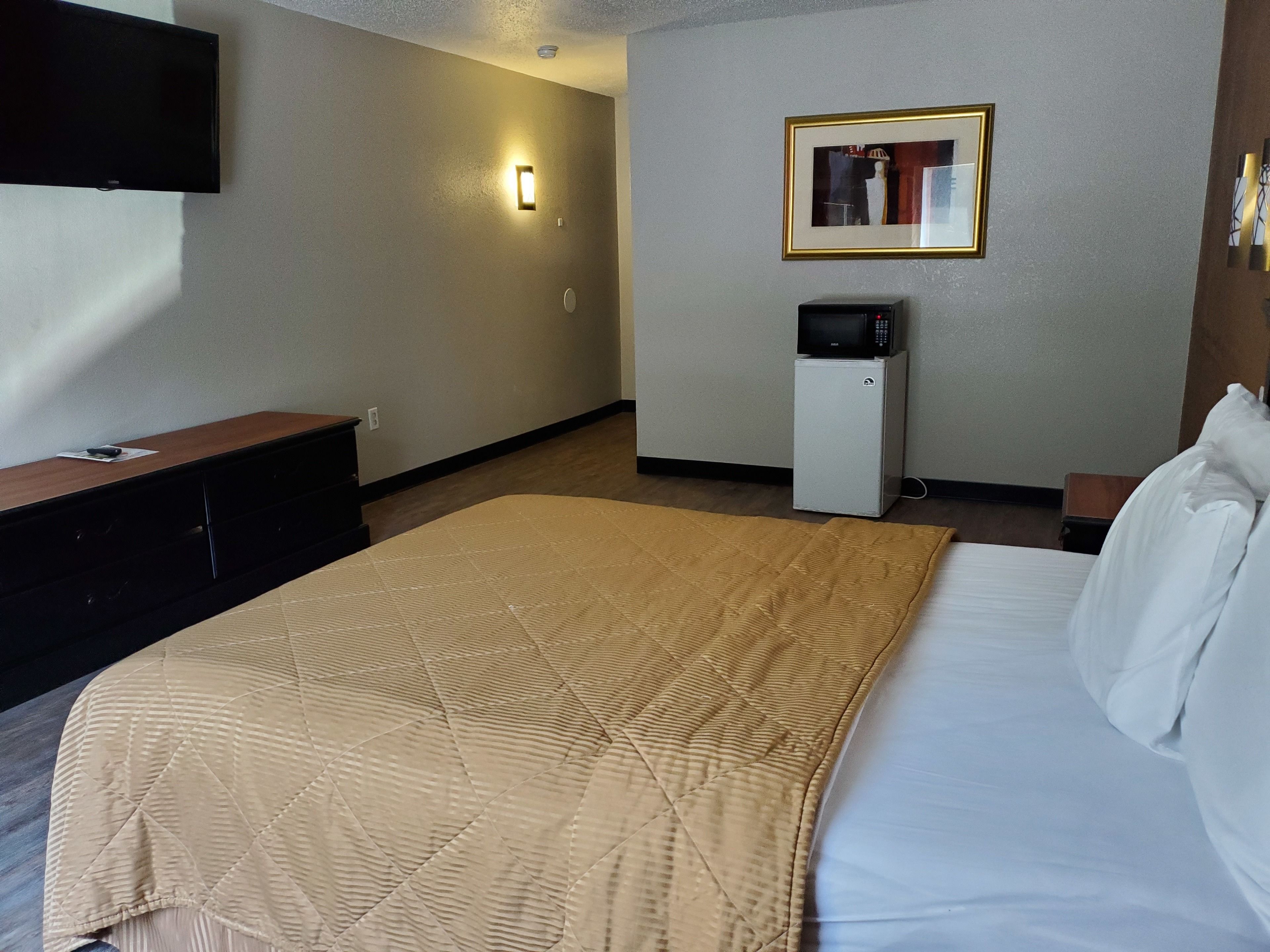 A hotel room with a bed, a TV mounted on the wall, a small refrigerator, and a framed picture on the wall.