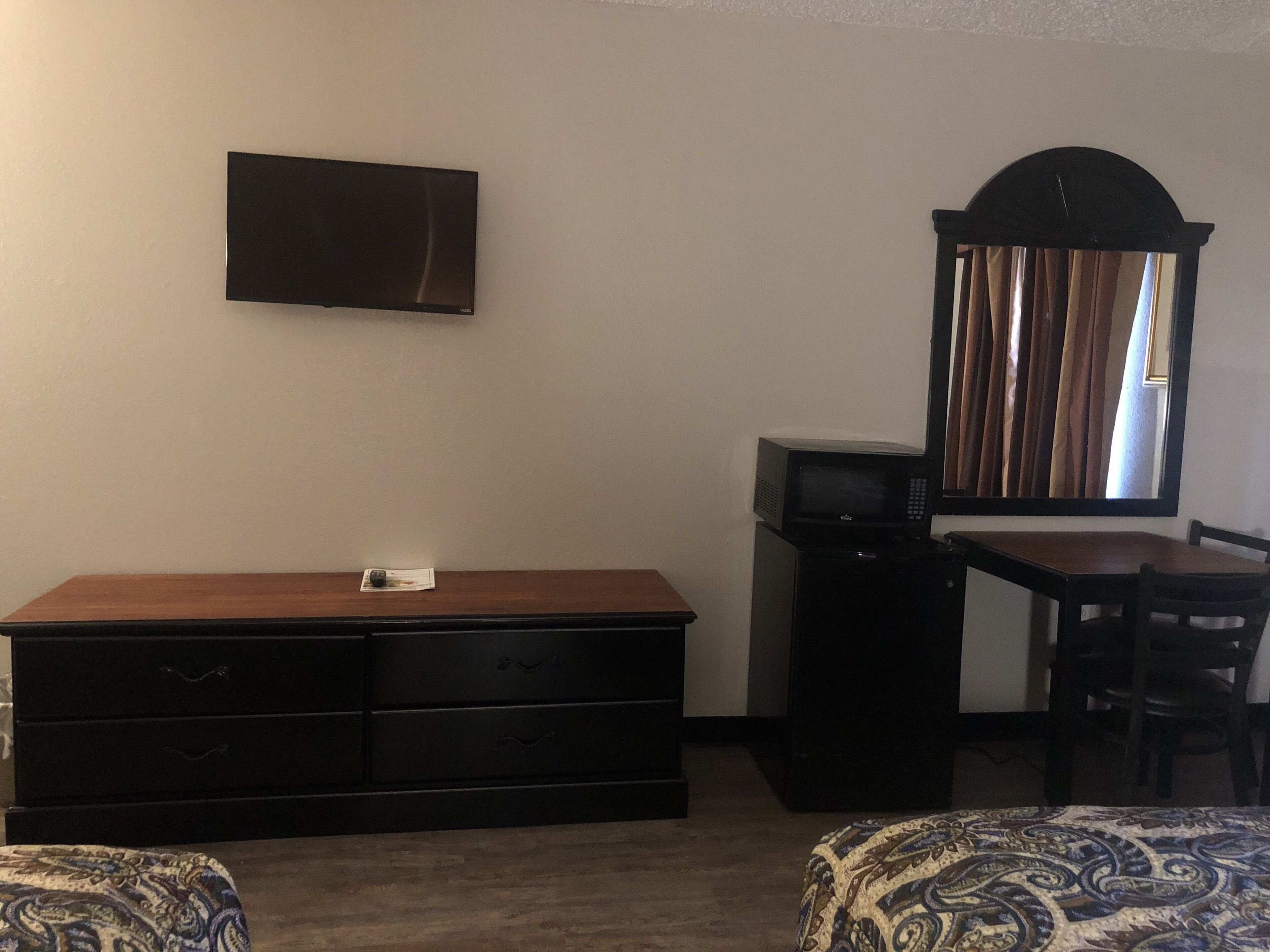 A room with a wooden dresser, a television mounted on the wall, a microwave, a dining table with chairs, and a patterned ottoman.