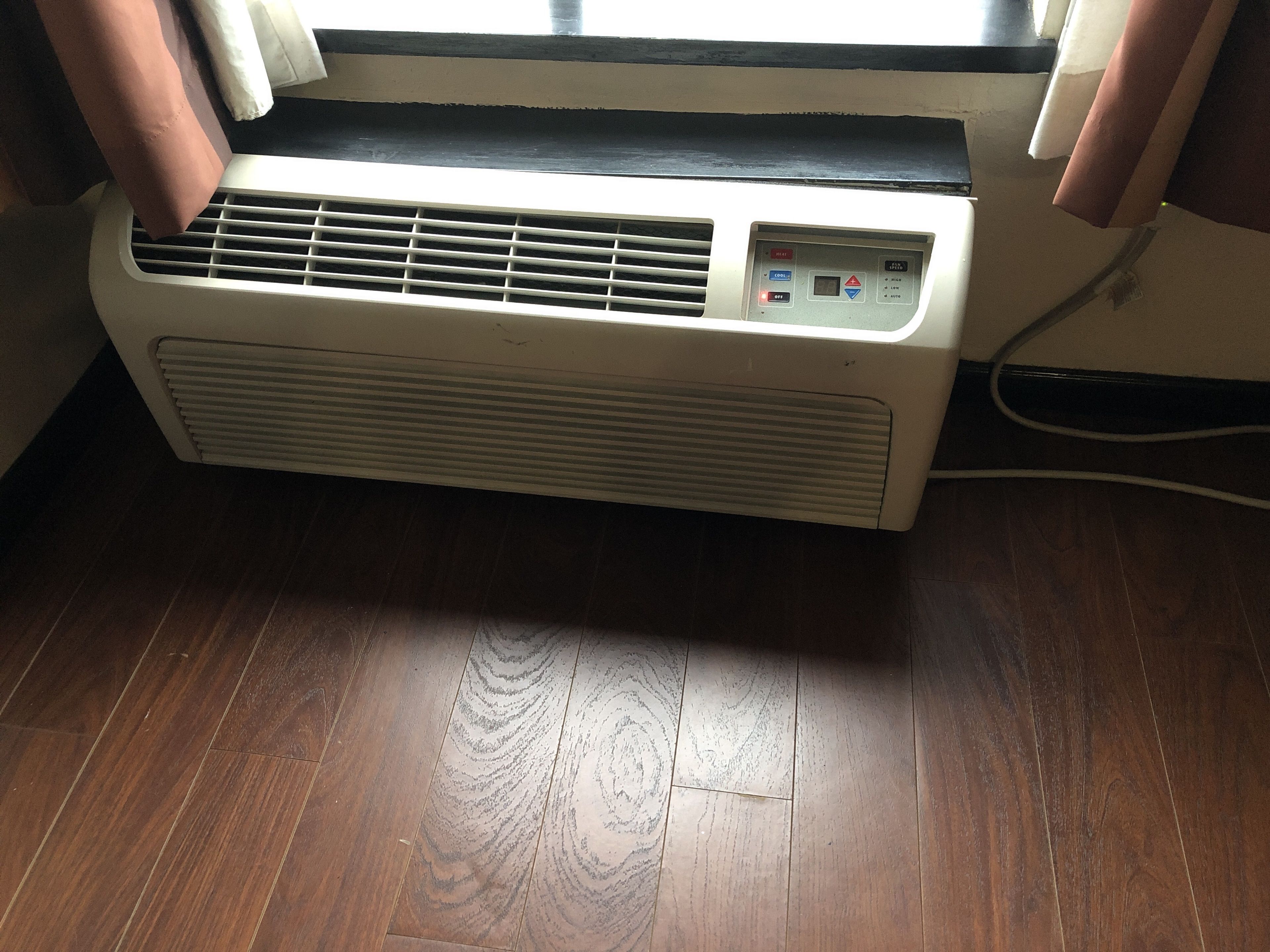 A portable air conditioner unit with a control panel, placed on a wooden floor near a window with curtains.