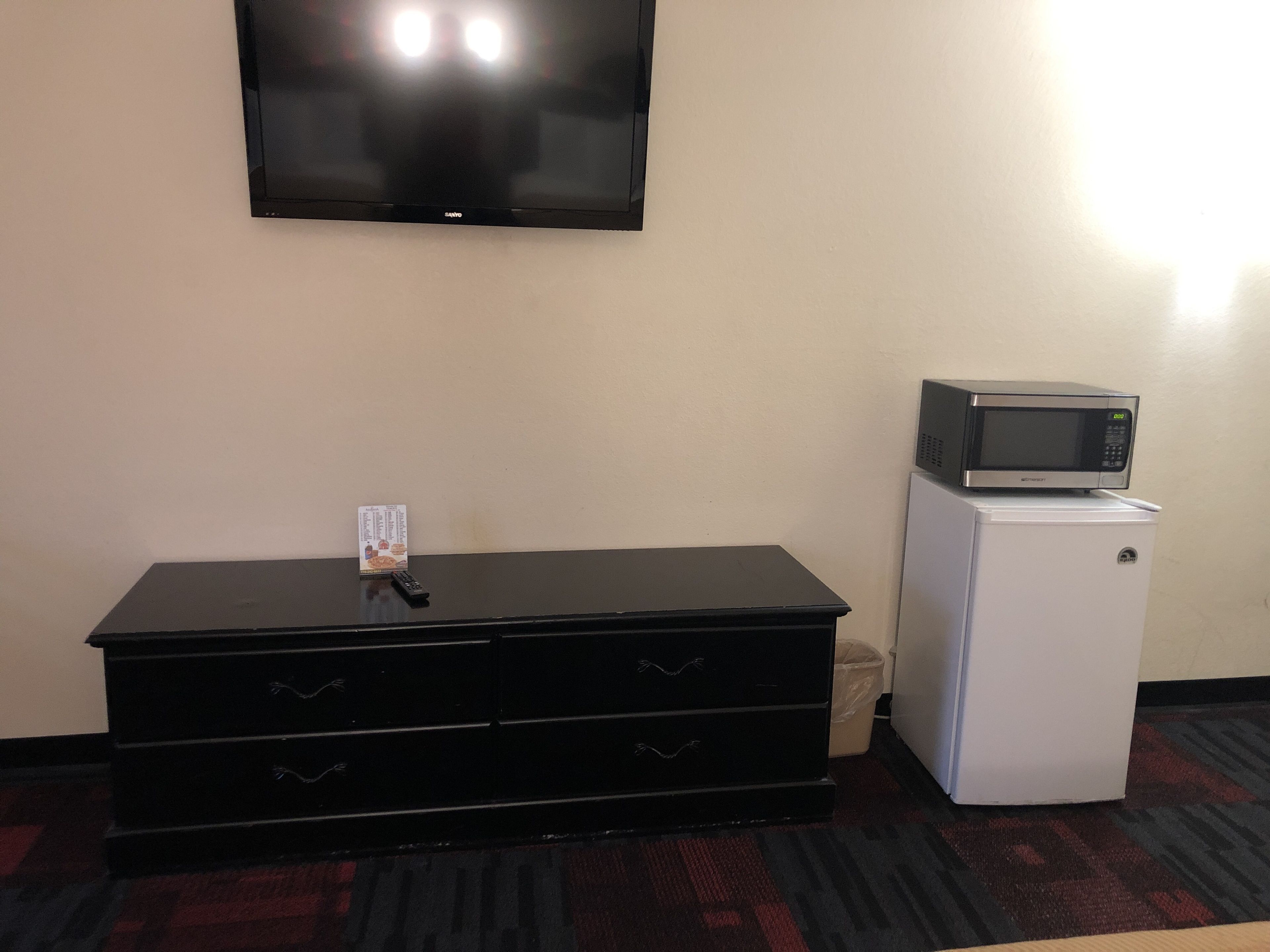 A room with a flat-screen TV mounted on the wall, a black dresser with drawers, a white refrigerator, and a microwave on top of the fridge.