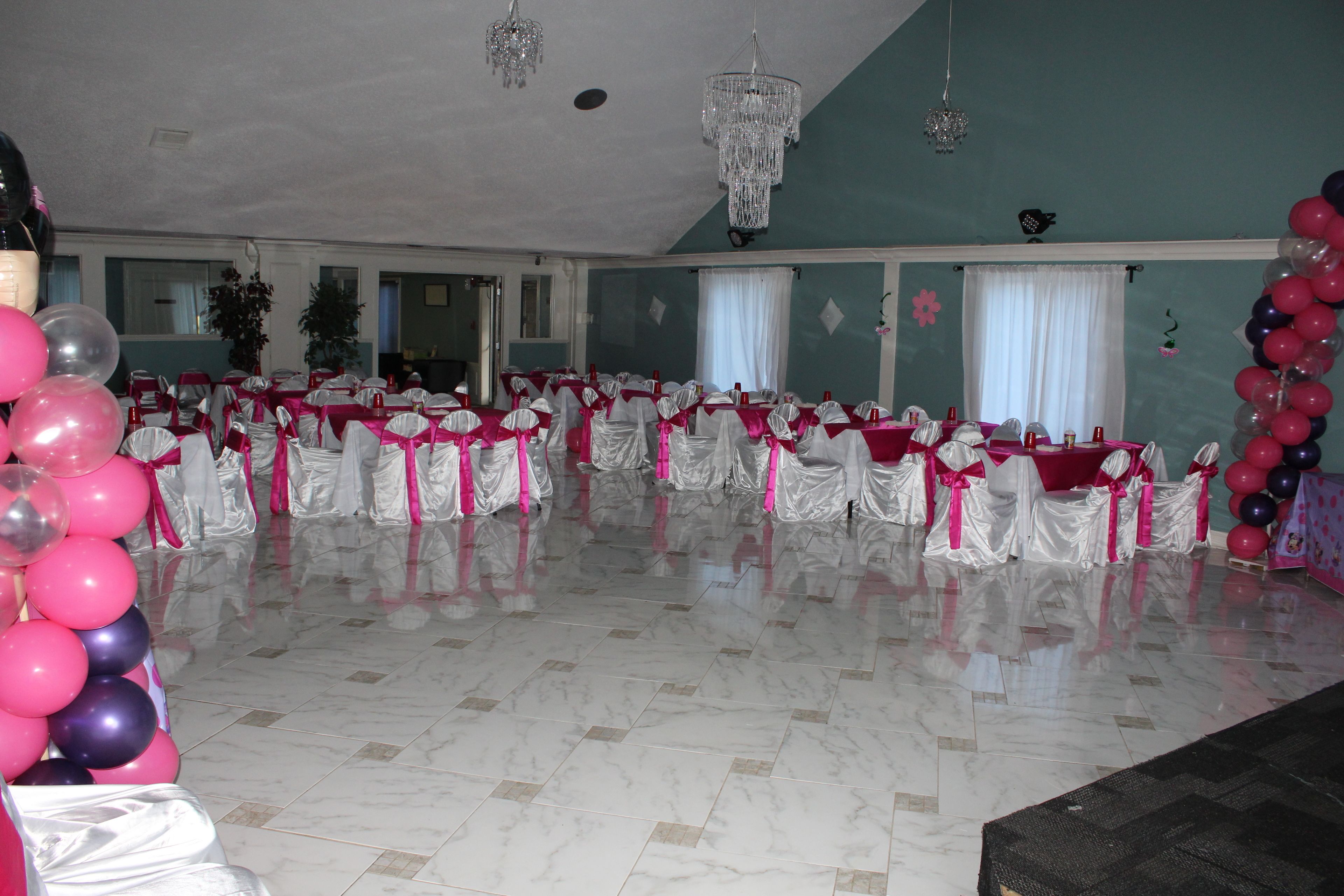 A spacious hall with rows of tables and chairs, chandeliers, and a large window with curtains.