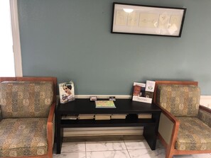 A seating area with patterned chairs, a small table with magazines and a framed picture, and a wall-mounted sign.