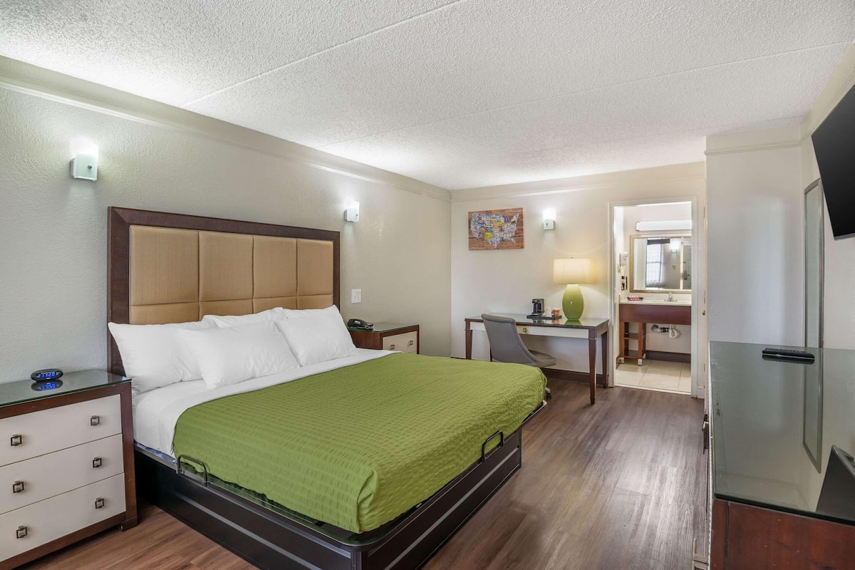 Habitación de hotel con cama, escritorio, silla, mesita, espejo y refrigerador.
