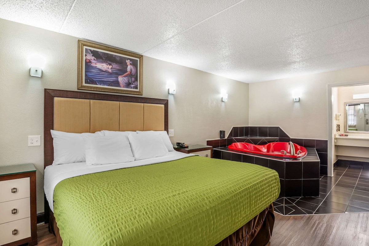 Habitación de hotel con cama, una bañera roja, mesita de noche y lámpara de pared.