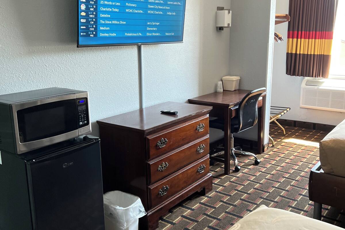 A hotel room with a television displaying a channel guide, a microwave, a refrigerator, a wooden dresser, a desk, a chair, and a bed.