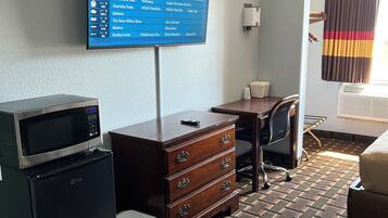 A hotel room with a television displaying a channel guide, a microwave, a refrigerator, a wooden dresser, a desk, a chair, and a bed.